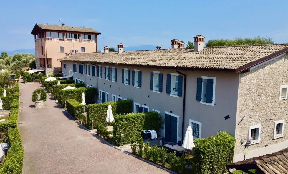 A row of beige, stone buildings with blue shutters surrounded by neat hedges and umbrellas in a garden courtyard at La Bertoletta Village apartments