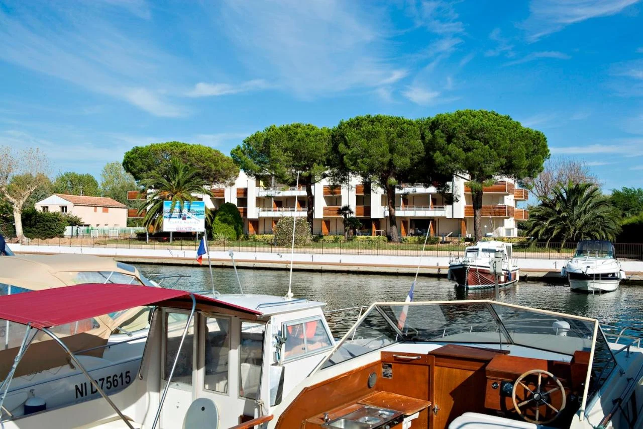 Boats are docked along a canal with a large, white residential building and lush green trees in the background at Residence Carré Marine