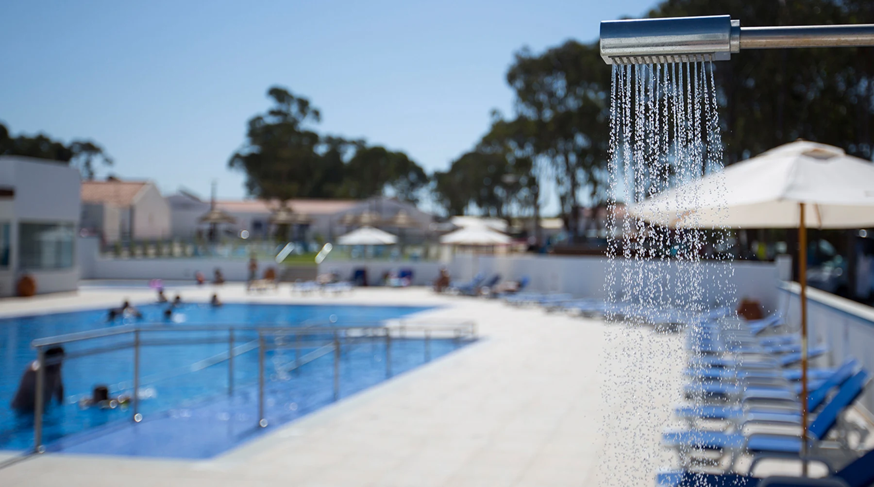 A poolside shower pours water near swimmers in a blue-tiled swimming pool, with lounge chairs and umbrellas in the background at Costa do Vizir Beach Village