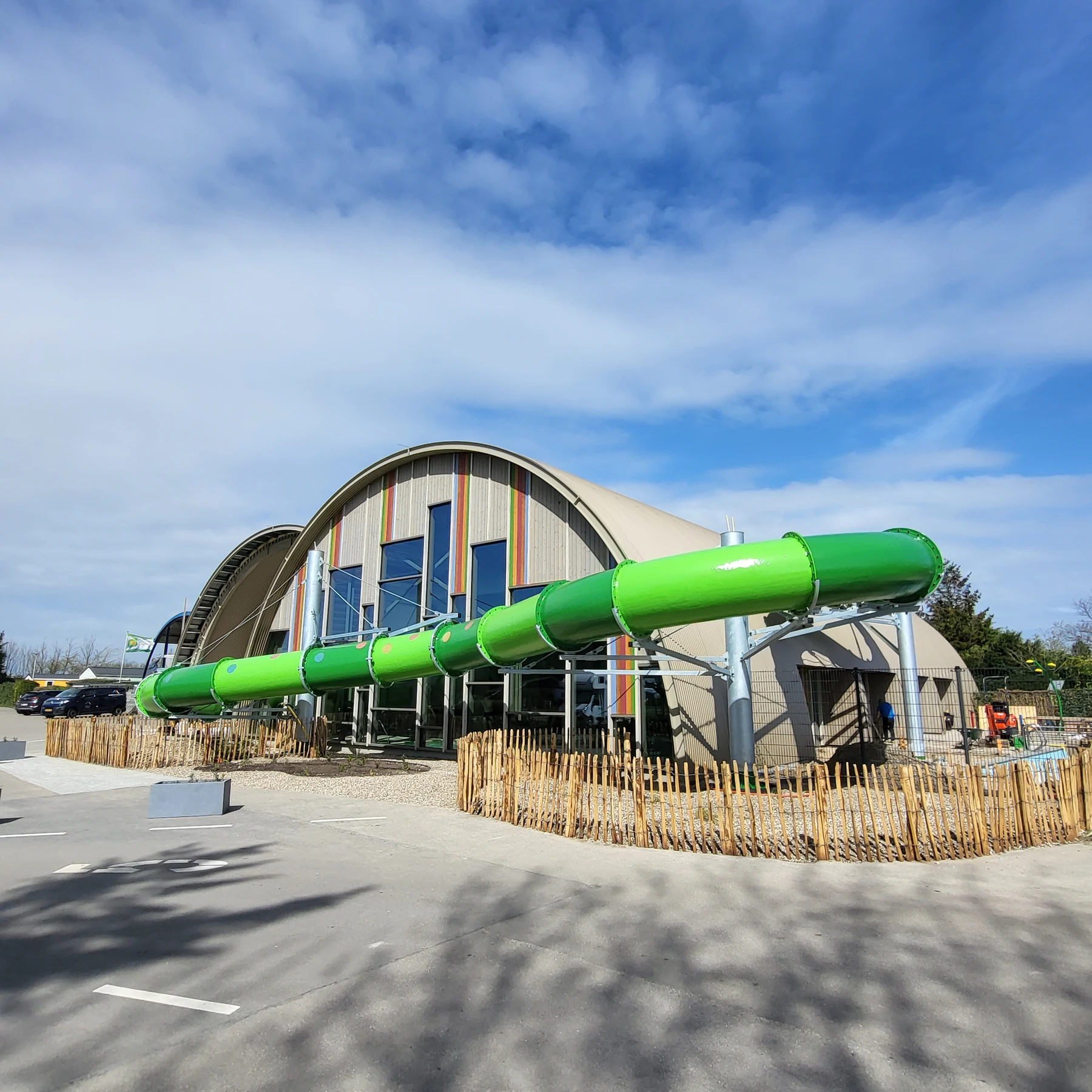 Green waterslide curves along a modern building with arched roof; parked cars and wooden fencing surround it at Vakantiepark Koningshof - Eurocamp