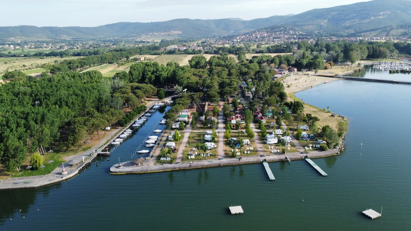 Boats docked at a marina, surrounded by trees, campsites, and distant mountains at Punta Navaccia