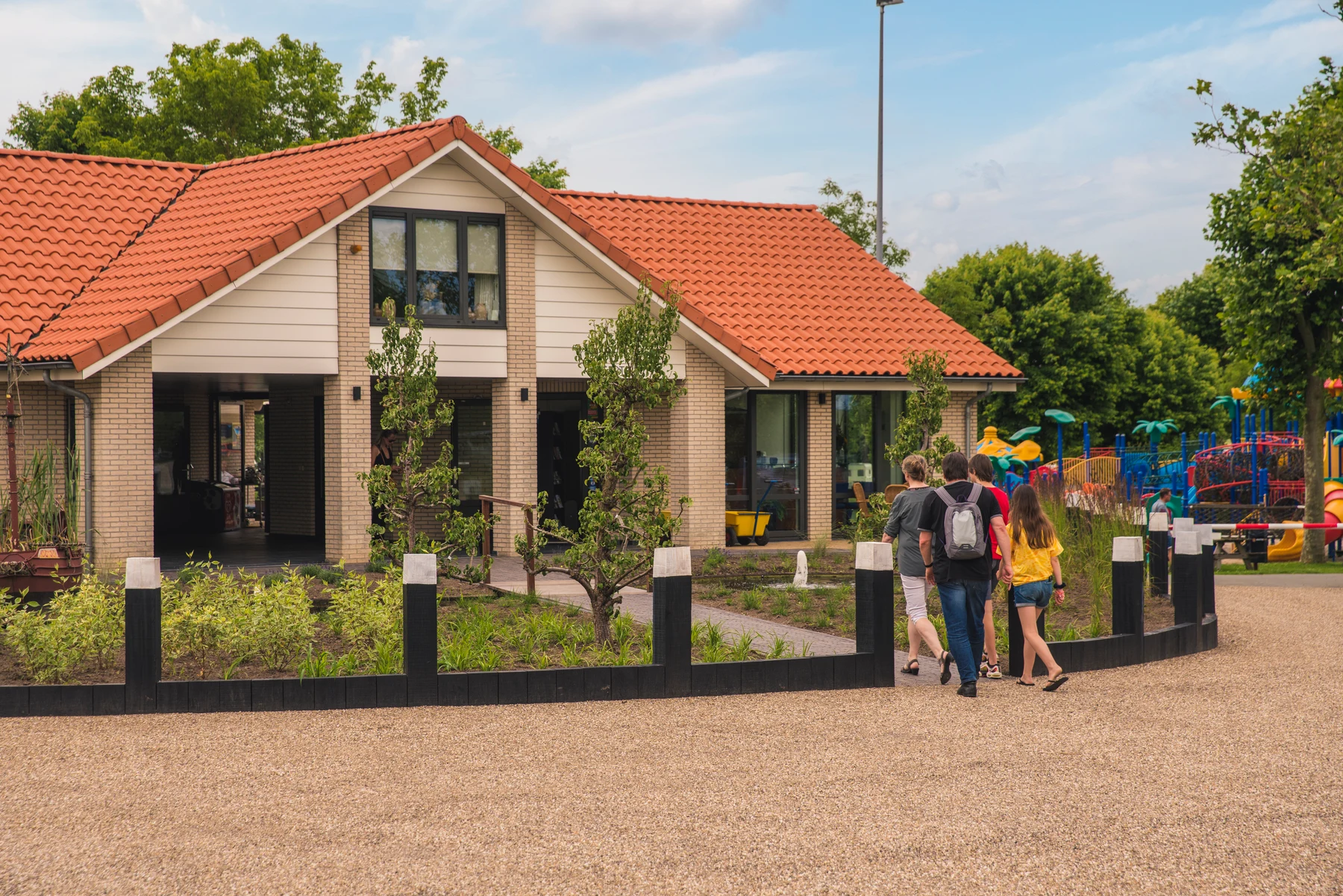 House with red-tiled roof, five people walking towards it, playground equipment and greenery surrounding at Marinapark Bad Nederrijn