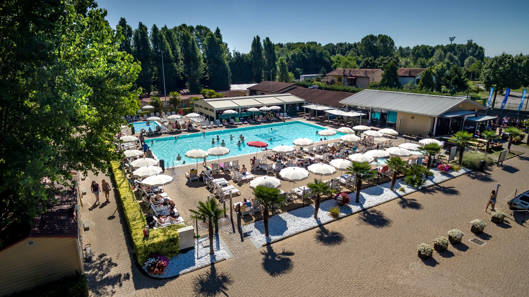 Swimming pool area with people lounging under umbrellas and swimming, surrounded by trees and buildings at hu Venezia Camping in Town