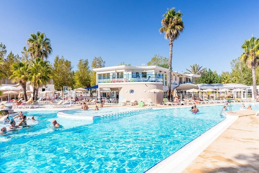 People swimming and sunbathing around a large outdoor pool with palm trees and a modern building in the background at Marvilla Parks Club Le Napoléon