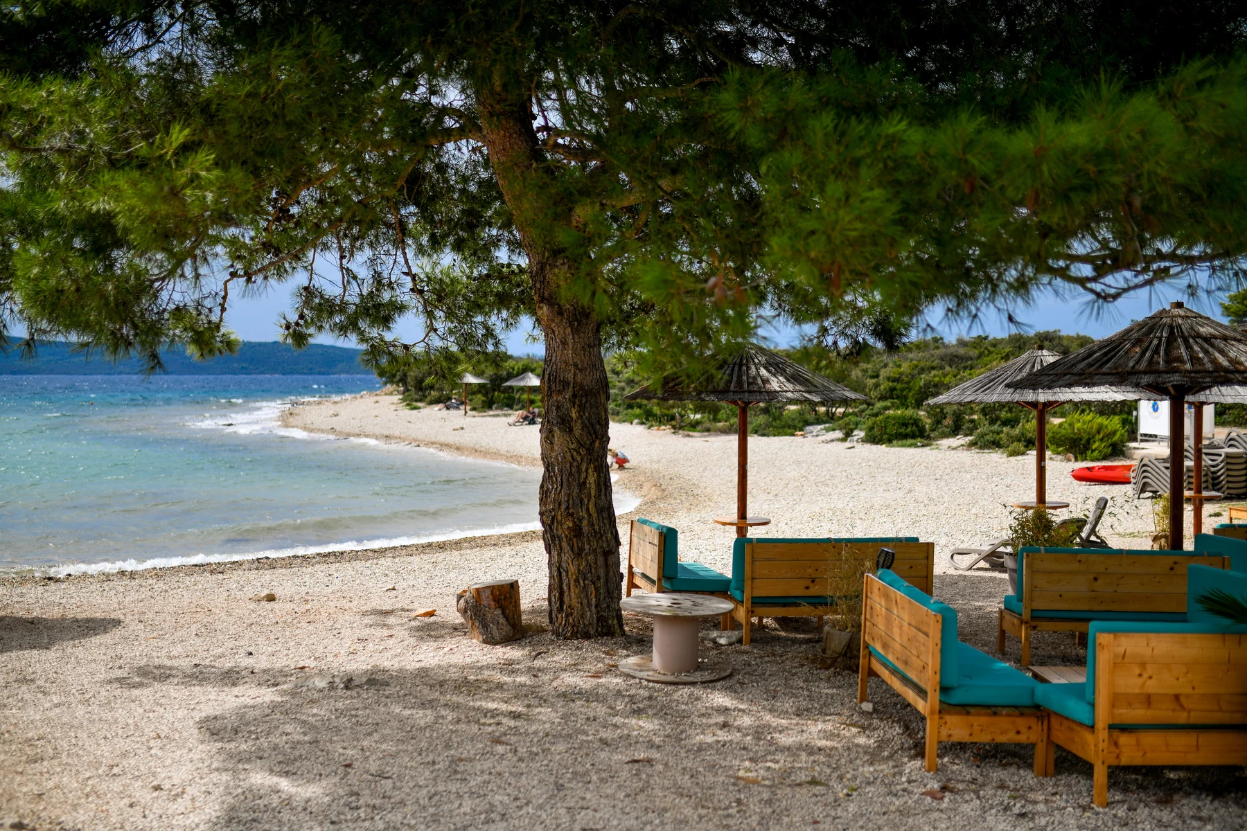 Wooden lounge chairs with blue cushions arranged under straw umbrellas on a pebbled beach near the sea at Ugljan