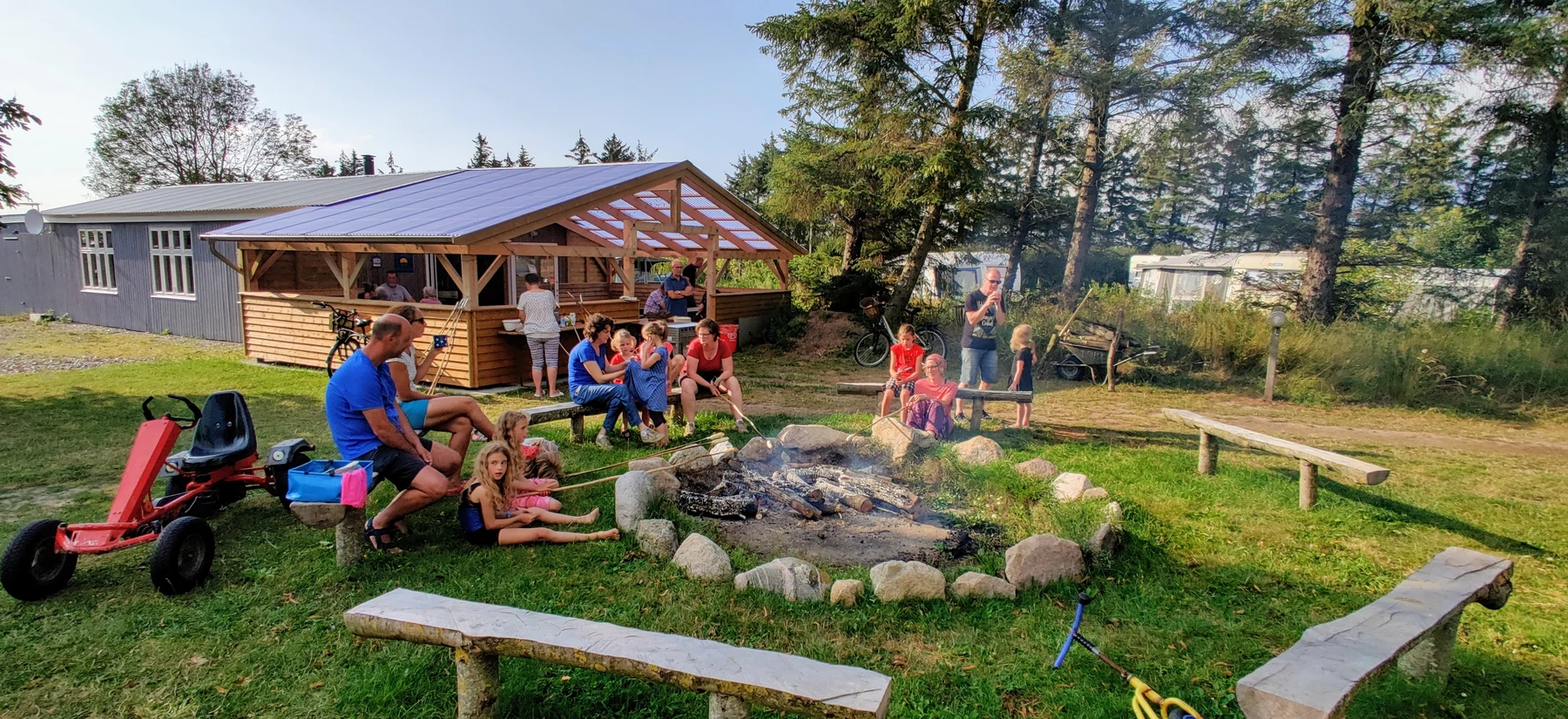 People sitting around a campfire roasting food, with wooden shelter and parked bikes in a forested campsite at Ballum