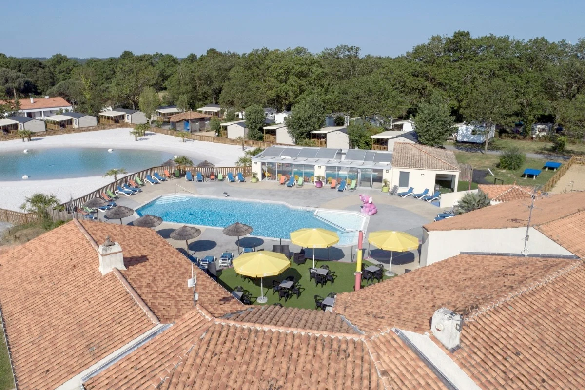 Swimming pool with yellow umbrellas and lounging chairs surrounded by cabins and a small lake within a forested campsite environment at Le Bois Joli