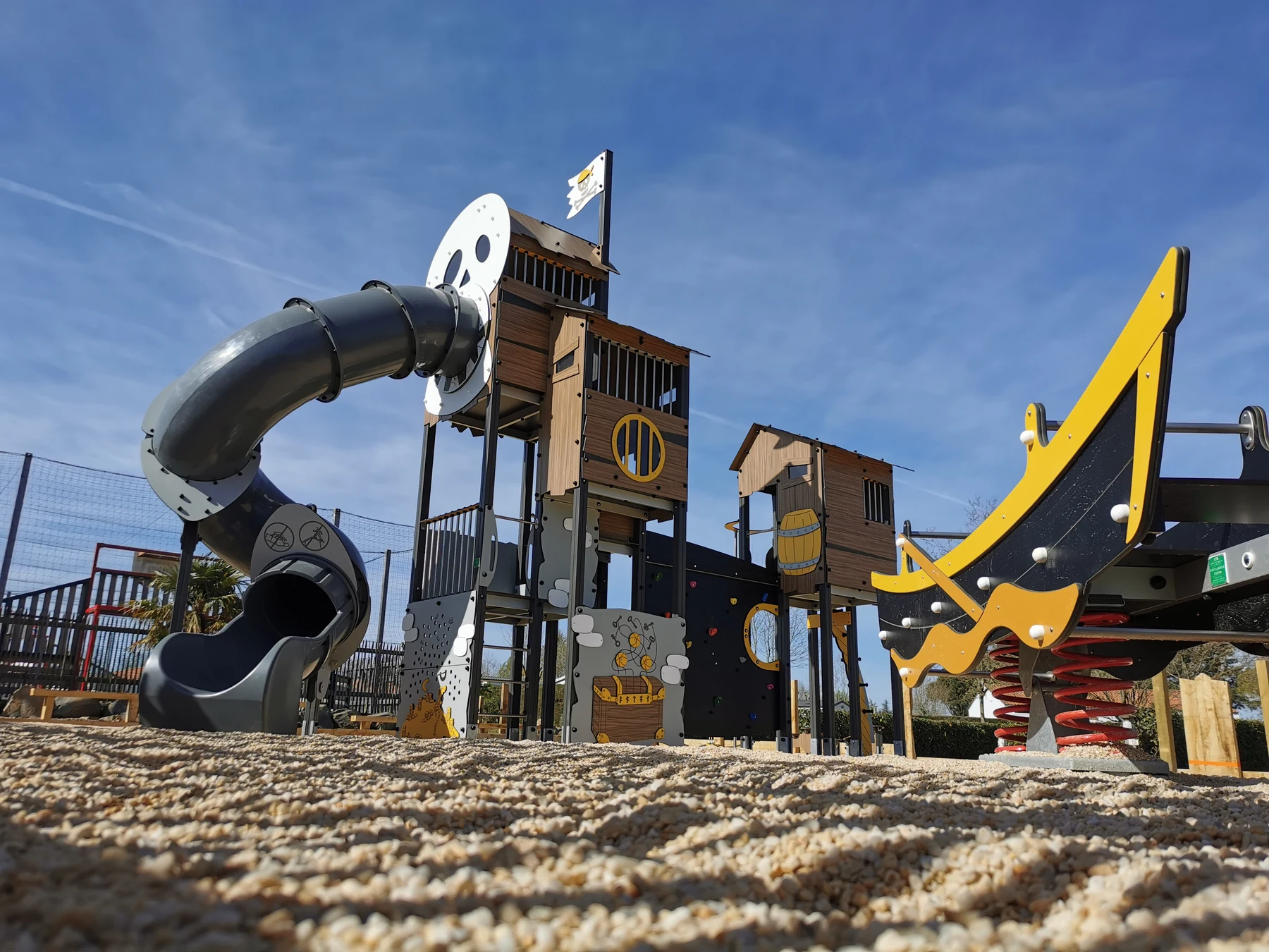 Pirate-themed playground set with slides and climbing elements on sandy ground under a clear, blue sky at La Bolée d'Air