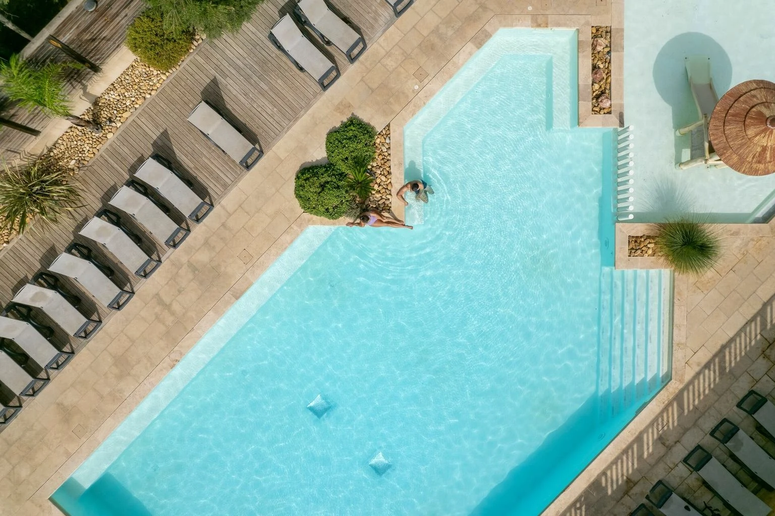 Two individuals relaxing at a swimming pool surrounded by lounge chairs and greenery at La Barque