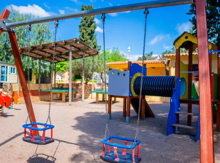 Swings hanging on a frame, adjacent to a colorful playground structure in a sunny outdoor area at Sant Salvador