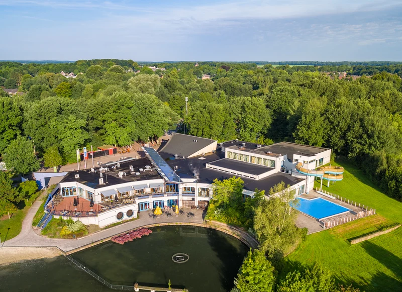 A large, modern building with a pool sits beside a pond, surrounded by lush trees at Hunzedal