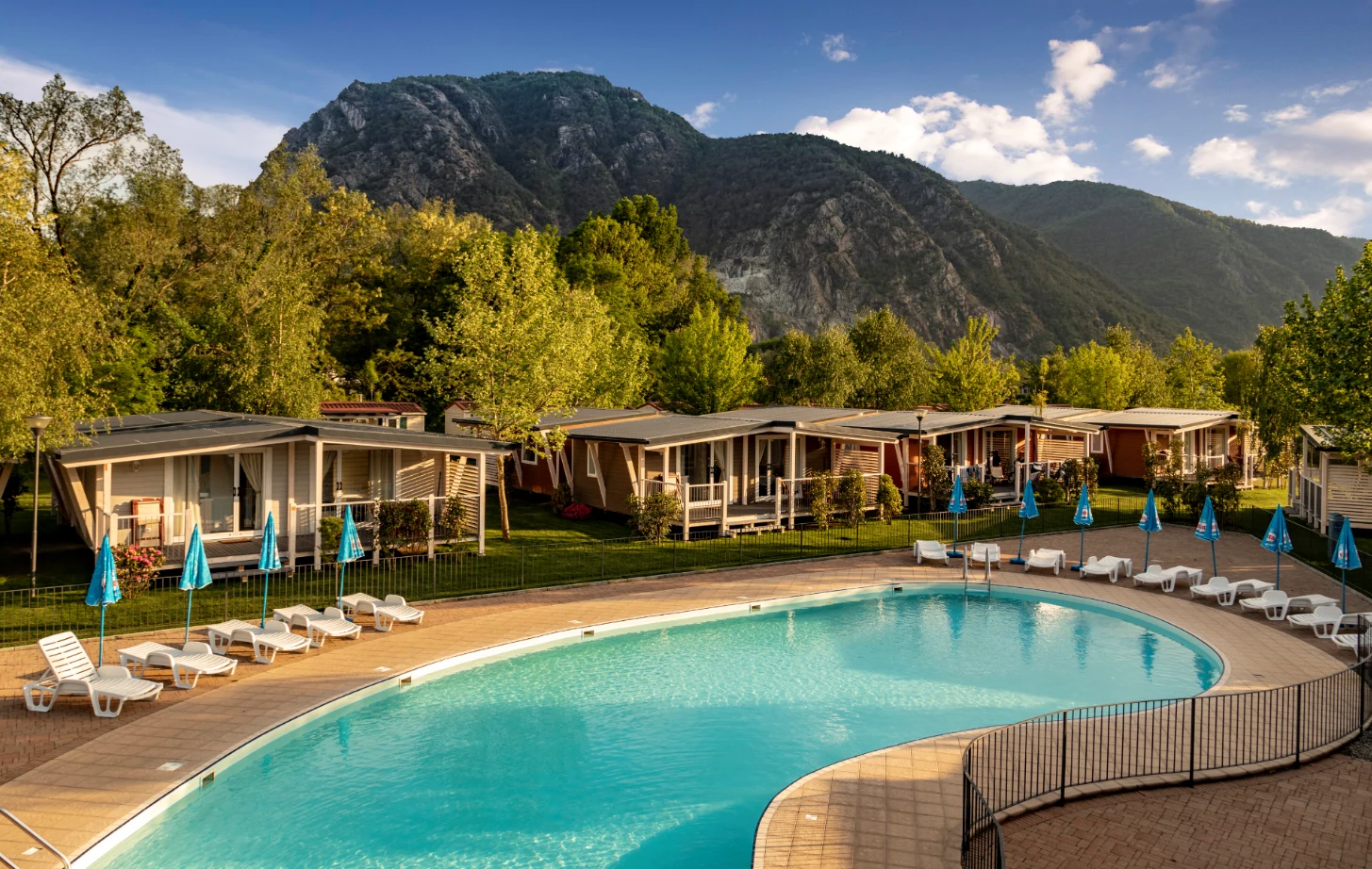 Swimming pool with sun loungers and umbrellas, surrounded by modern cabins, nestled in a scenic mountainous area at Glamping II Borgo delle Arti