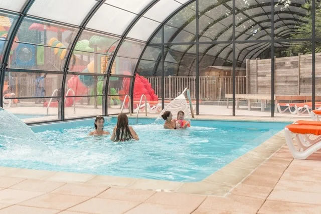 Children swimming indoors; enclosed pool with splashing water, colorful play structures, and lounge chairs around at Château de Rochetaillée