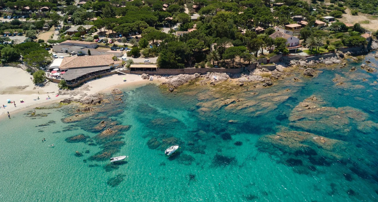 Boats float on clear turquoise water near rocky shoreline and sandy beach, surrounded by trees and buildings at U Livanti