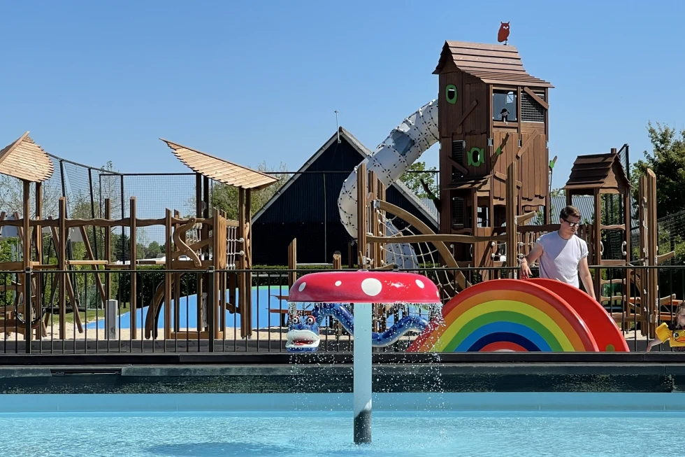 Playground with tall wooden structures, slides, and a colorful rainbow sculpture beside a water spray feature at Camping de Molenhoek
