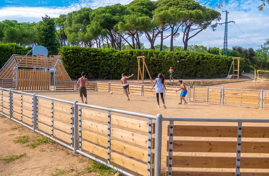 Children playing soccer in a fenced dirt field, surrounded by trees and playground structures at 3 Estrellas Costa 