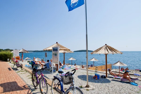 Bicycles parked beside sunbathing people under straw umbrellas near a pebble beach by the sea at Zelena Laguna