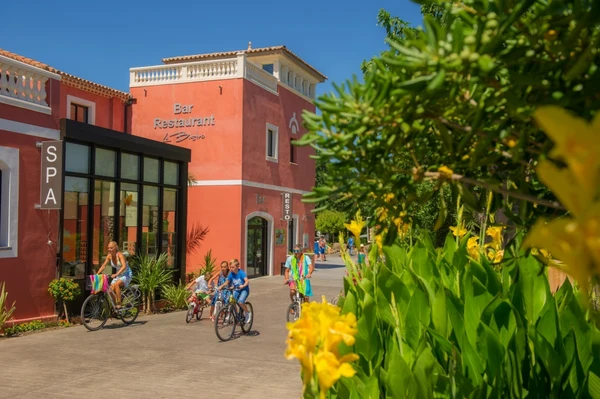 People ride bicycles along a path beside a red building with a restaurant and spa, amidst vibrant green foliage at Les Méditerranées Beach Garden