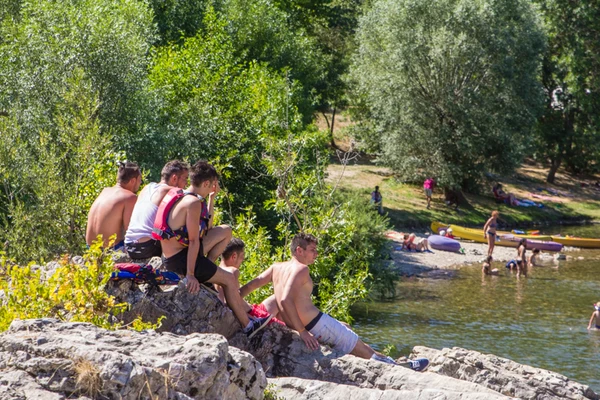 Group of people sitting on rocks, observing swimmers and kayaks by a riverside, surrounded by trees at du Domaine d'Anglas
