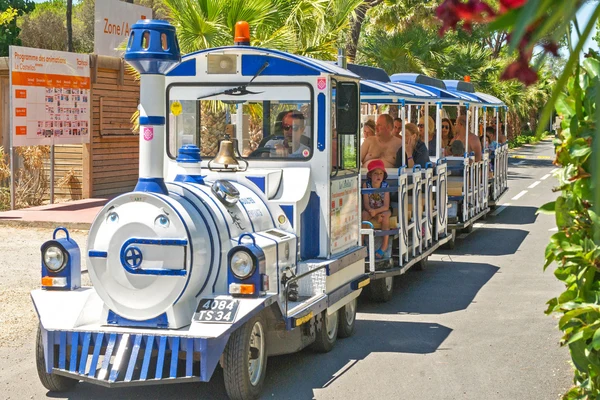 Miniature train carrying people, travels on paved road surrounded by trees and posters at Marvilla Parks Le Castellas