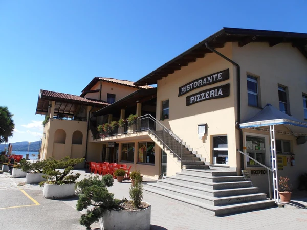 Restaurant building with a staircase, outdoor seating, and potted plants under clear skies; located at Lido di Monvalle