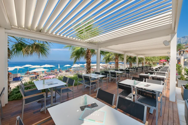 Tables and chairs are arranged under a pergola, overlooking a beachfront with palm trees and umbrellas at Luxury homes Medora Orbis