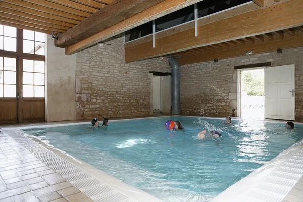 Children swimming in an indoor pool with wooden beams and stone walls at Château de l'Epervière