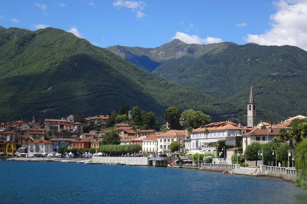 Town buildings line a waterfront, backed by lush green mountains under a blue sky at Glamping II Borgo delle Arti