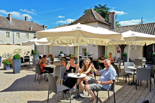 People sitting, drinking, and enjoying under large umbrellas in an outdoor cafe courtyard with stone buildings in the background at Château de l'Epervière