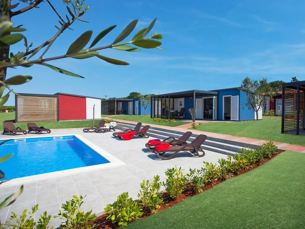 A swimming pool with lounge chairs, surrounded by colorful modern cabins, sunny day, green lawn at Kastanija