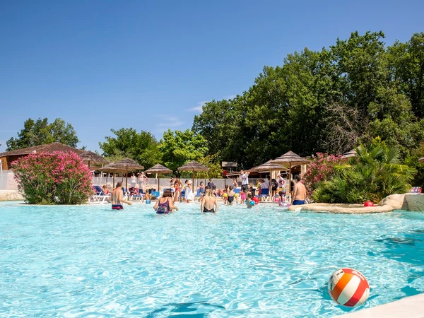 People swimming and relaxing in a large outdoor pool surrounded by trees, flowers, and sun umbrellas at l'Évasion