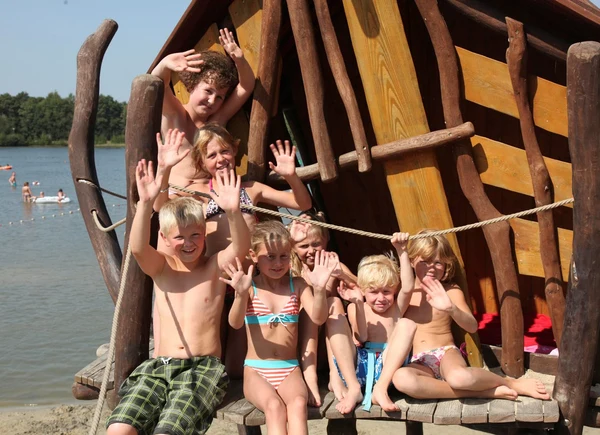 Children waving and smiling on wooden play structure beside lake with people swimming in the background at Hunzedal