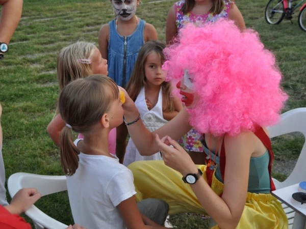 Clown with pink wig paints child's face while other children watch outdoors at San Francesco