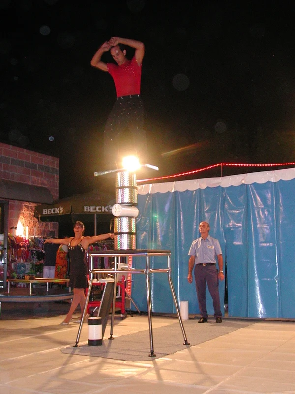 Performer balances on stacked cylindrical objects under a spotlight, with two onlookers nearby at Village Torre Pendente