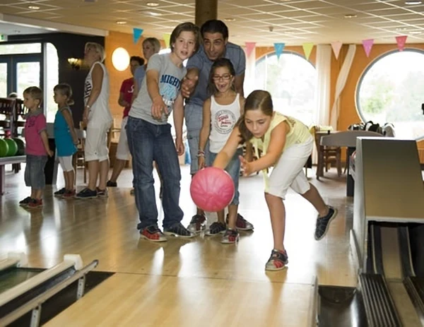 A girl is bowling with guidance from a man, watched by children indoors with colorful flags at Hunzedal