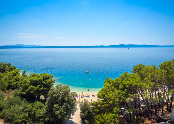 People relaxing on a sandy beach surrounded by trees and overlooking a calm, blue sea with distant mountains at Luxury homes Medora Orbis