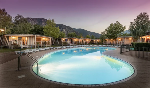 Swimming pool reflecting evening lights, framed by lounge chairs and modern cabins, with mountains in the background at Glamping II Borgo delle Arti