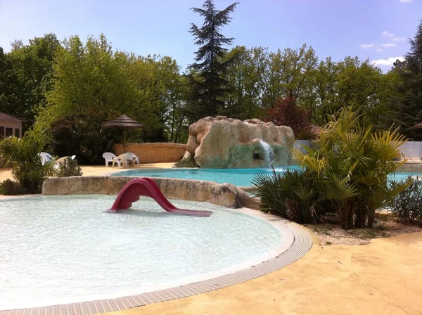 Red slide enters shallow pool near waterfall and lounge chairs in lush green outdoor area at l'Évasion