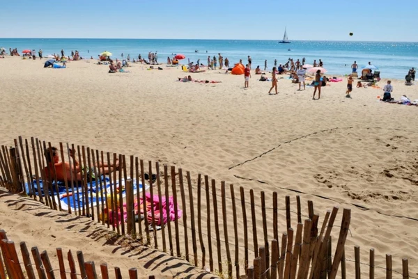 People sunbathing, playing, and relaxing on a sandy beach with the sea and a sailboat in the background at Les Mediterranées Nouvelle Floride