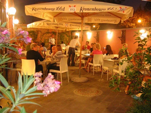 People dining under umbrellas, enjoying a meal on a well-lit outdoor patio surrounded by plants at Village Torre Pendente