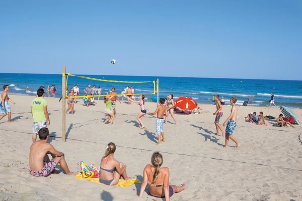 People playing beach volleyball, various spectators relaxing on the sand, near a vibrant blue ocean at Les Méditerranées Beach Garden