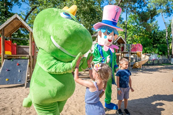 A child high-fives a green dinosaur mascot near playground structures with another mascot and children nearby at Flower Camping Les Biches