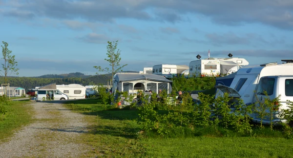 Caravans parked, people seated and relaxing, amidst trees and grass under a blue sky at Brilon
