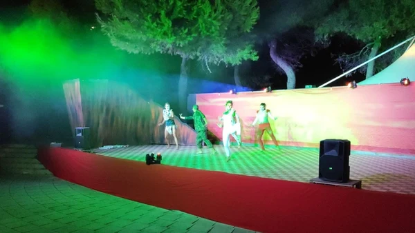Performers dance energetically under colorful lights, surrounded by trees on an outdoor stage at Savoy Beach Hotel & Thermal Spa