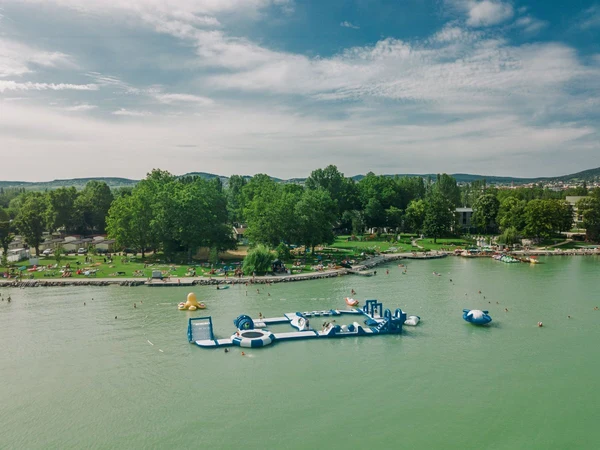 Inflatable water obstacle course floating in a lake, surrounded by people swimming and relaxing in a green park at Füred