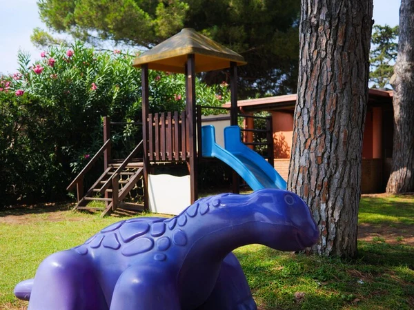 Purple dinosaur playground toy sits on grass near slide and tree, surrounded by greenery and buildings at Club Degli Amici