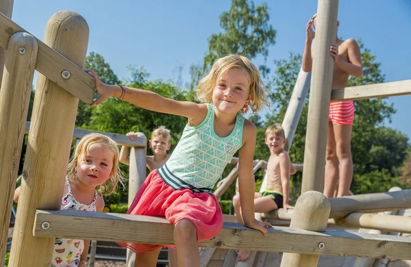 Children climbing wooden playground structure, enjoying sunny weather with lush greenery in the background at Hoeve de Schaaf