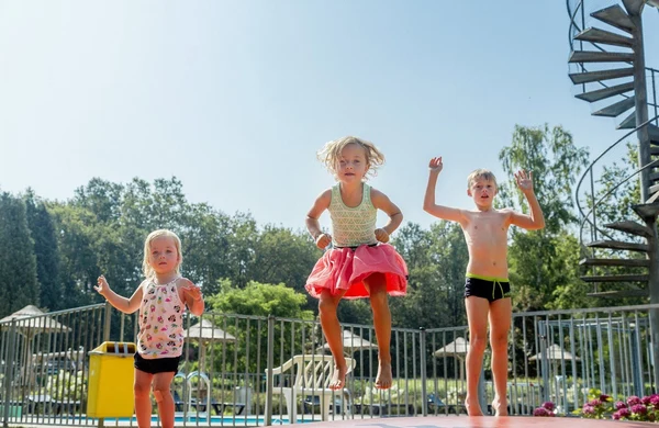 Three children jumping excitedly on a trampoline, surrounded by green trees and a spiral staircase at Hoeve de Schaaf