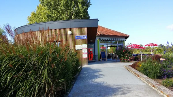 Building labeled "Reception," surrounded by lush plants and colorful flowers, with outdoor seating under red umbrellas at Seasonova Du Chêne