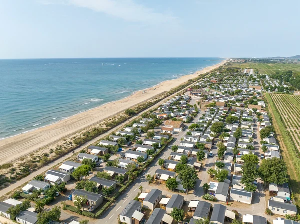 Rows of mobile homes line a coastal campsite, adjacent to a sandy beach and ocean, under a clear sky at Marvilla Parks Le Castellas